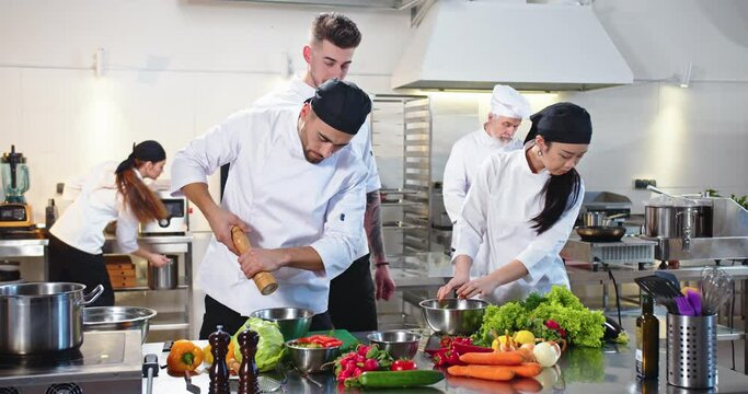 Workflow in cafeteria kitchen. Busy mixed-race team of chefs preparing food in famous restaurant. Caucasian young man cook preparing meal in restaurant with Asian woman colleague, culinary concept