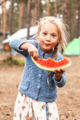 A child, a girl, with blond hair smiles against the backdrop of a camping, eating a watermelon, a portrait to the waist, shooting in full face