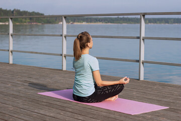 A young caucasian pretty mindful woman meditates holding fingers in yoga sign sitting on a mat on the sea pier.Peaceful girl keeps hands in mudra gesture sitting in the lotus position.Morning, sunrise