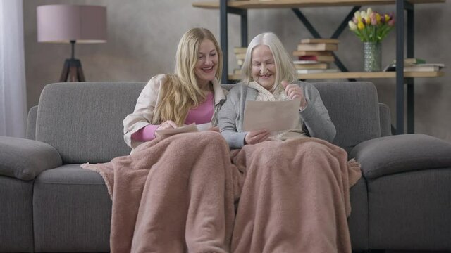 Wide shot front view of joyful happy retirees looking at pictures talking and laughing. Portrait of relaxed cheerful Caucasian women recalling good memories sitting on couch in living room
