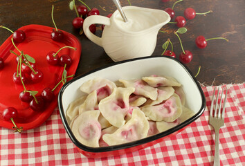 Dumplings with cherries in a red container next to a milk jug with sour cream and cherries on the table. Cooked dumplings with cherries for lunch, recipe. Russian cuisine