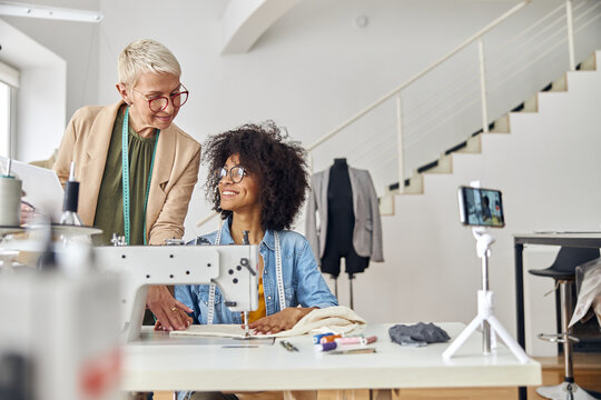 Middle aged woman helps young African-American colleague to sew in fashion studio