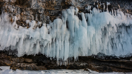 Fancy icicles on the slope of a sheer granite rock. Close-up. Details. Ice lace over a frozen lake. The rough surface of the stones. Baikal in winter