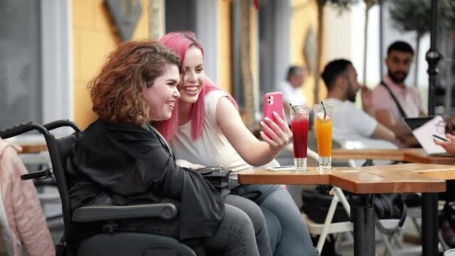A happy disabled woman is taking a selfie photo with her friend while sitting on the street cafe outside