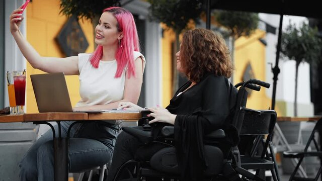 A pretty nice disabled woman is taking selfie photo while having a business lunch with her friend sitting on the street cafe outside