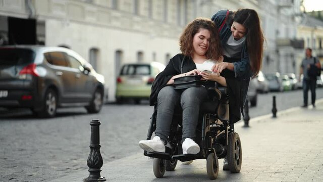 A pleased disabled woman is spending time with her friend is walking and using her smartphone on the city street