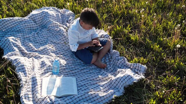 The boy is sitting in a field with a tablet in his hands. The child will scroll on the computer. The boy is playing. Outdoor recreation. Beautiful sunset. The planet is in danger. Healthy lifestyle