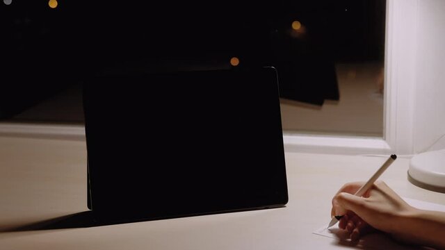 Distance education. Online test. E-learning school. Video conference. Unrecognizable woman writing on paper with portable computer blank screen night home interior copy space.