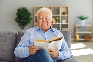 Smiling elderly man or grandfather in blue shirt sitting on sofa and reading book at home