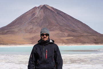 Young man with sunglasses and hut stands in front of a turquoise lagoon and a dry mountain in the Andean highlands of Bolivia.