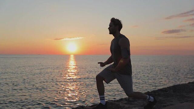 Strong sporty athletic toned fit sportsman man 20s wear sports clothes warm up training doing stretch exercise for legs at sunrise sun dawn over sea beach outdoor on pier seaside in summer day morning