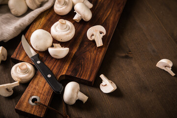 Fresh raw white Champignon mushrooms on dark brown moody wooden cutting board with kitchen knife and linen towel