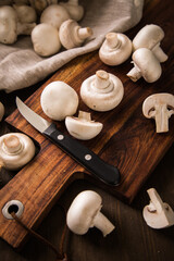 Fresh raw white Champignon mushrooms on dark brown moody wooden cutting board with kitchen knife and linen towel