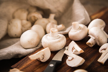 Fresh raw white Champignon mushrooms on dark brown moody wooden cutting board with kitchen knife and linen towel
