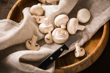 Fresh raw white Champignon mushrooms on linen towel with kitchen knife, wooden bowl and dark brown moody background