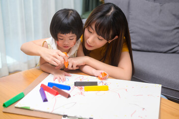 Young mom helping daughter drawing with colored pencils in living room at home.