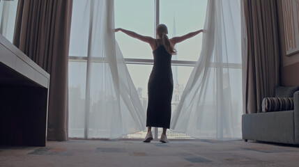 Back view of woman opening curtains and looking out of window to cityscape at hotel in the morning. Modern buildings at the Dubai city