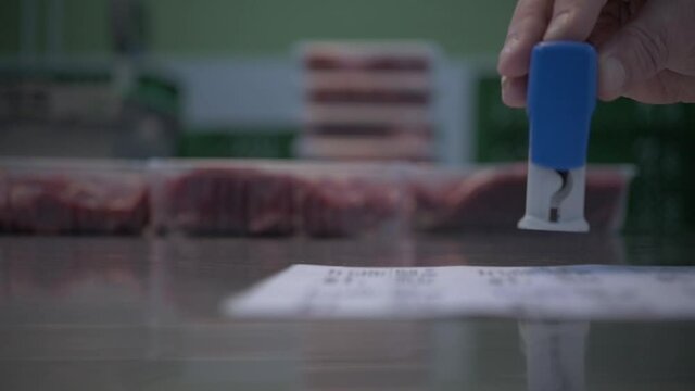Hands of worker puts stamp on the labels for marking trays with different cuts of fresh raw beef meat on table in slaughterhouse. Slow motion. Meat production and food industry,