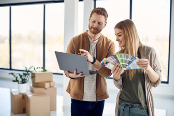 Smiling couple using laptop while choosing color from palette during home renovation.
