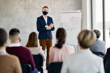 Businessman with face mask talking to large group of colleagues on presentation at conference hall.