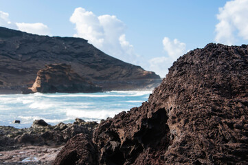 Fototapeta na wymiar Kanaren, Lanzarote,. Küstenlinie mit typischen schwarzem erkaltetem Lavafelsen und kräftiger Brandung. Im Hintergrund die typische vulkanische Landschaft