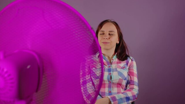 Close up of young woman sitting on chair in front of fan on purple background. Happy brunette with closed eyes enjoying of ventilator air.