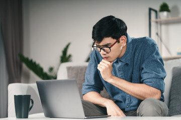 Pensive Thoughtful Serious Asian man in eyeglasses looking at laptop screen thinking for solution to solve the problem..