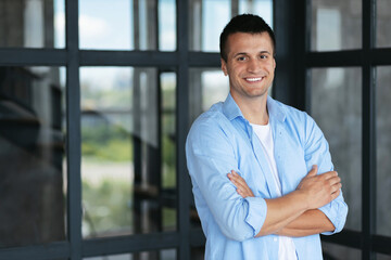 Successful young caucasian businessman with arms crossed standing in the office, smiling, looks at the camera