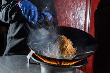 Funchoza rice noodles with vegetables cooking on fire in wok pan. Street flambe food
