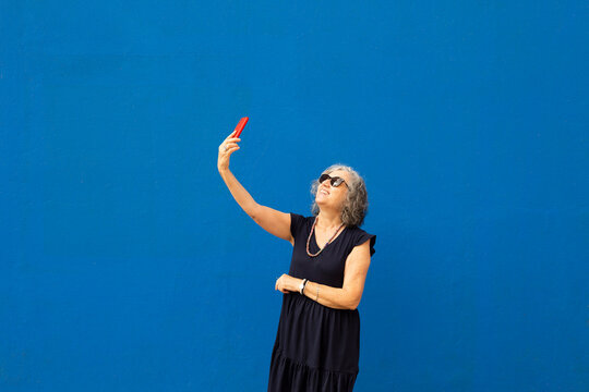 Senior grey haired woman taking a selfie with a red smartphone against a blue wall