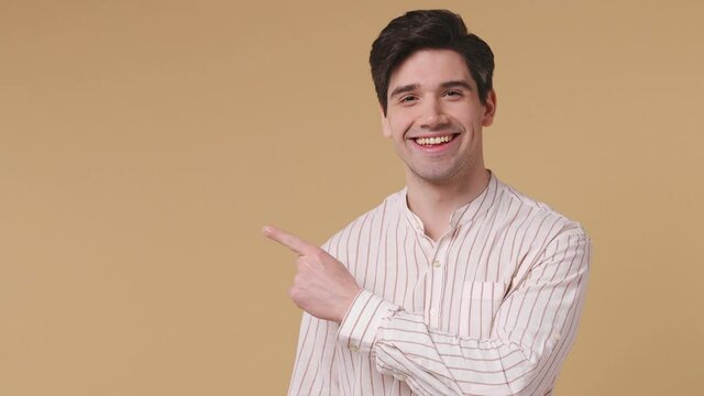 Happy young promoter man 20s years old wears white striped shirt pointing fingers hands aside on workspace copy space mockup promo commercial area isolated on pastel beige background studio portrait