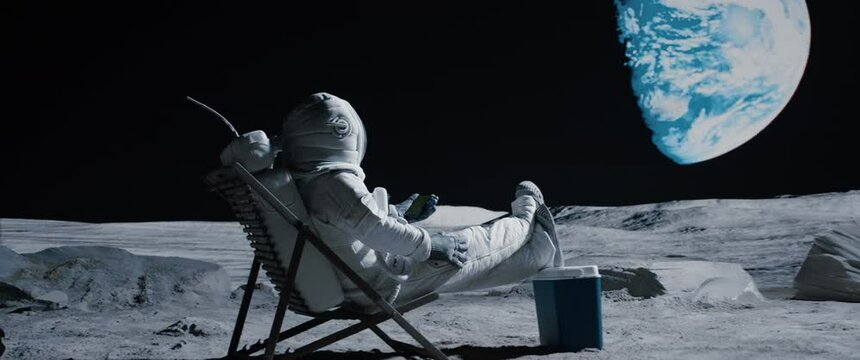 Astronaut sits in a beach chair on a Moon surface, holding phone in hands. Shot with 2x anamorphic lens