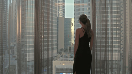 Silhouette of young woman opens curtains on the big window stretches arms and looking out her apartment on the city buildings during amazing sunrise. Dubai city