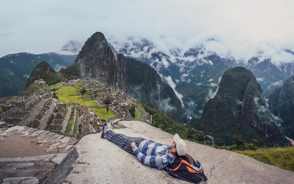 Panoramic shot of young man laying down, contemplating Machu Picchu lost city with Huayna Picchu. Ruins of ancient inca civilization in the sacred valley of Cusco Province. Peru, South America