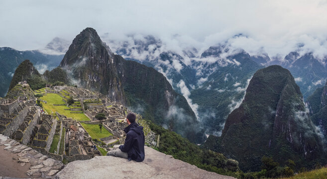 Panoramic shot of young man sitting, contemplating Machu Picchu lost city with Huayna Picchu mountain. Ruins of ancient inca civilization in the sacred valley of Cusco Province. Peru, South America