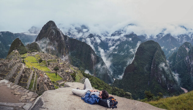 Panoramic shot of young man laying down, contemplating Machu Picchu lost city with Huayna Picchu. Ruins of ancient inca civilization in the sacred valley of Cusco Province. Peru, South America