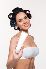 smiling young woman in curlers and bra showing cream tube isolated on white.