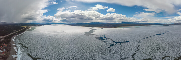 Fototapeta na wymiar Panoramic drone aerial view of a frozen lake breaking up in northern Canada during spring time with large ice landscape on blue sky day. Marsh Lake, Yukon.