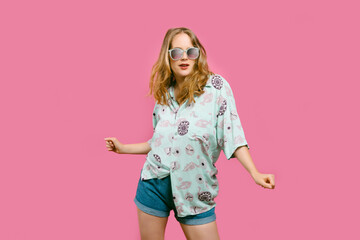 Portrait of a young blonde girl in a hippie shirt and sunglasses dancing on a pink background. summer concept and music festival. copy space