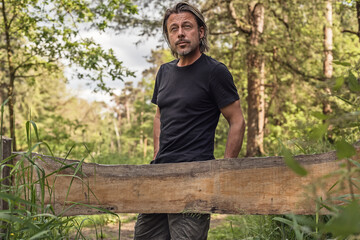 Man with stubble beard and blond hair in a black t-shirt stands by a wooden fence in a forest. © ysbrandcosijn