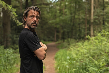 Blond man in a black t-shirt with crossed arms on a forest path.