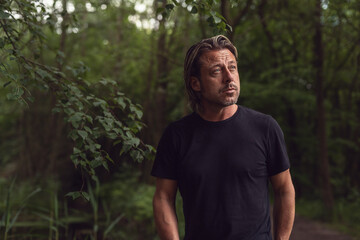 Blond man with stubble beard in a black t-shirt in a lush forest.