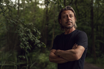 Blond man with crossed arms and stubble beard in a black t-shirt in a lush forest.