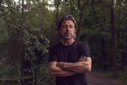 Blond man with crossed arms and stubble beard in a black t-shirt in a lush forest.