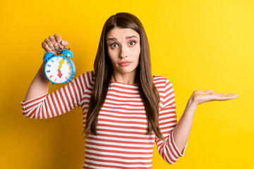 Portrait of attractive puzzled clueless girl holding clock shrugging shoulders isolated over bright yellow color background