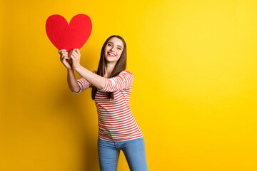 Portrait of attractive cheerful girl holding big large heart card having fun isolated over bright yellow color background