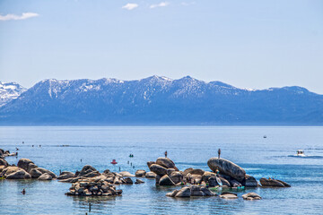 Tourists in water on paddleboards and climbing on rocks in blue Lake Tahoe with snow tipped...