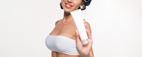 partial view of young woman in curlers and top showing cream tube isolated on white, banner.