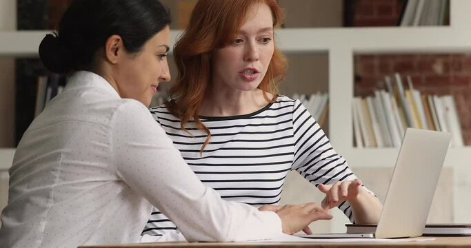 Happy young indian ethnicity team leader helping concentrated red-haired female colleague with online project on computer. Two mixed race millennial employees developing company growth strategy.