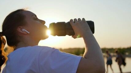 Fototapeta na wymiar Young woman drinks water from a bottle after a workout. Jogging outdoors in rays of the sunset. Outdoor sports in the park in summer, spring, girl drinks clear cool mineral water after training.